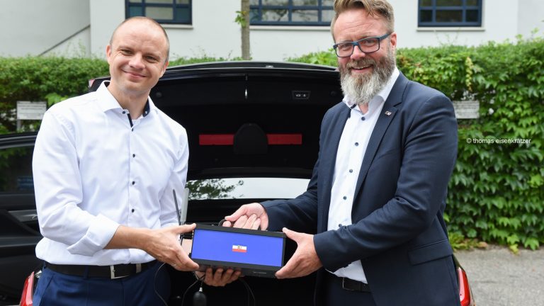 Zwei Männer stehen vor einem geöffneten Kofferraum. Beide tragen Hemden mit langen Ärmeln. Sie halten gemeinsam ein Gerät. Im Hintergrund sind eine Hecke und ein Gebäude mit Fenstern zu sehen.