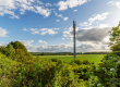 Ein Mobilfunkmast steht auf einem Feld unter einem Himmel mit verstreuten weißen Wolken. Umgeben von grünen Büschen und Bäumen erstreckt sich die Landschaft in die Ferne.