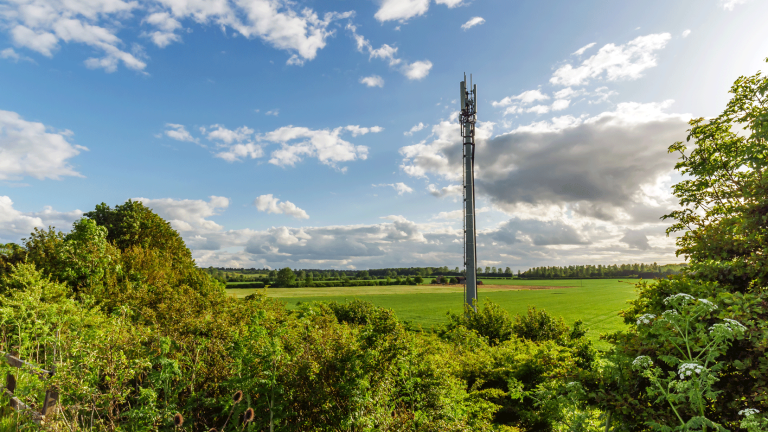 Ein Mobilfunkmast steht auf einem Feld unter einem Himmel mit verstreuten weißen Wolken. Umgeben von grünen Büschen und Bäumen erstreckt sich die Landschaft in die Ferne.