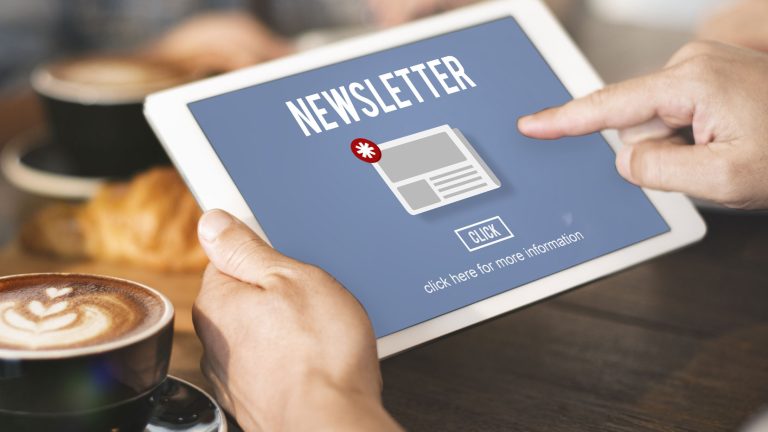 Eine Person hält ein Tablet in der Hand. Auf dem Bildschirm steht "Newsletter" mit einem Symbol für eine Benachrichtigung. Darunter befindet sich ein Button mit der Aufschrift "Click" und einem Hinweis zur weiteren Information. Im Hintergrund sind unscharfe Objekte.
