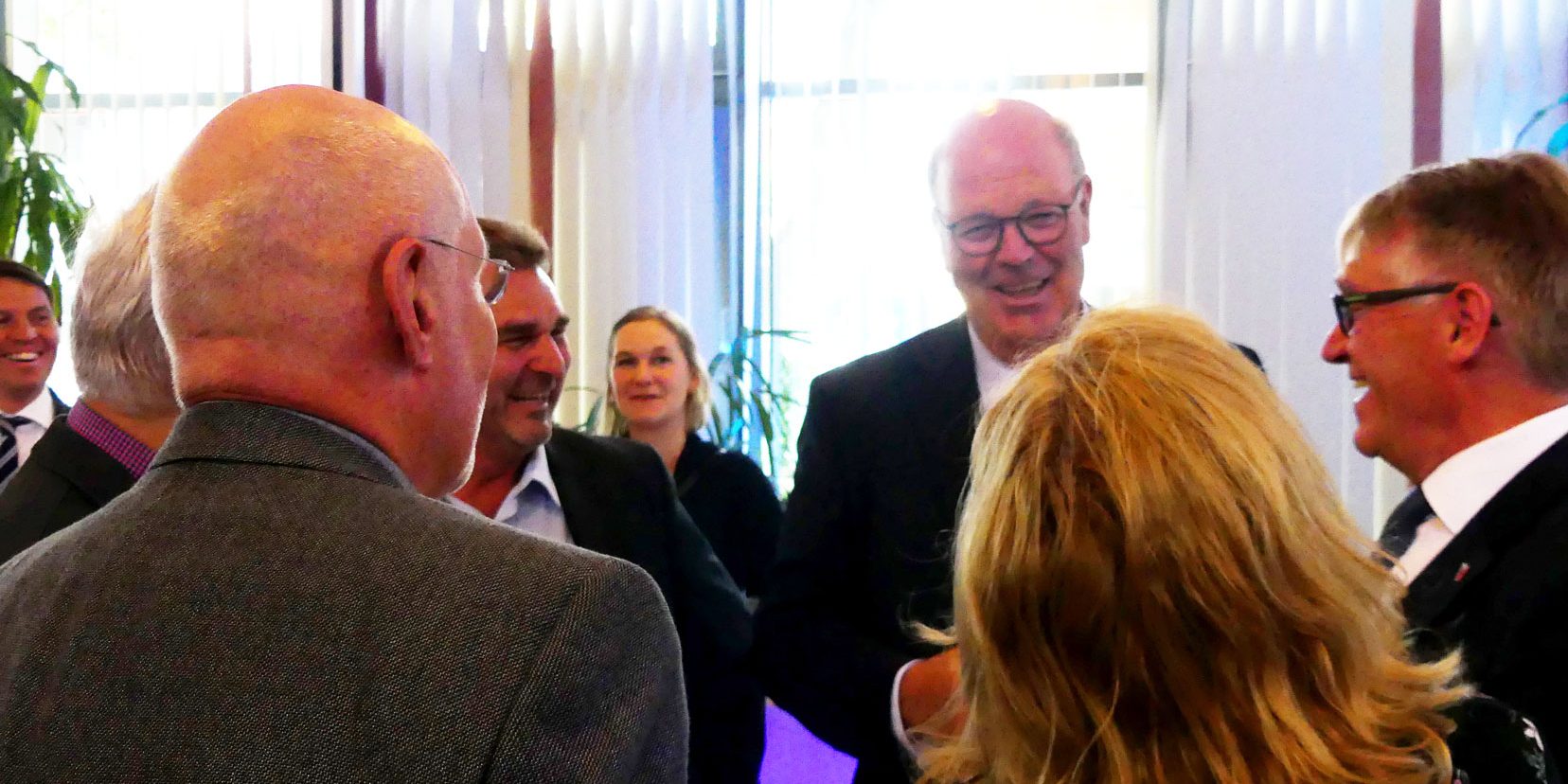 P1000984 Web Eine Gruppe von Menschen steht in einem hell erleuchteten Raum um einen Tisch und unterhält sich. Die Personen sind gut gekleidet, und es herrscht eine freundliche Atmosphäre. Im Hintergrund sind Fenster mit vertikalen Jalousien zu sehen.