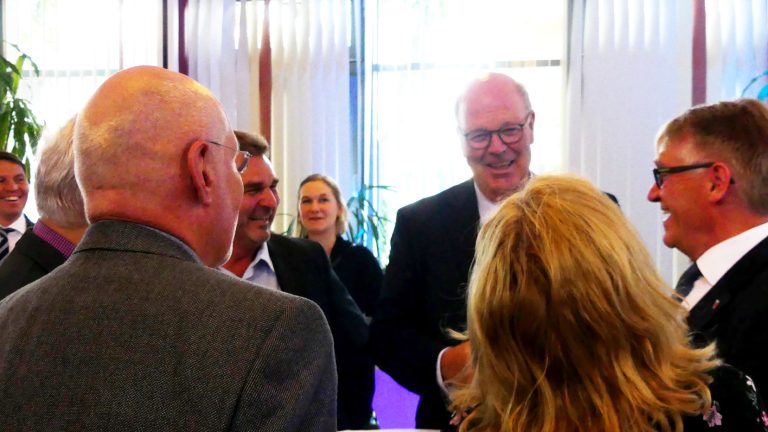 Eine Gruppe von Menschen steht in einem hell erleuchteten Raum um einen Tisch und unterhält sich. Die Personen sind gut gekleidet, und es herrscht eine freundliche Atmosphäre. Im Hintergrund sind Fenster mit vertikalen Jalousien zu sehen.