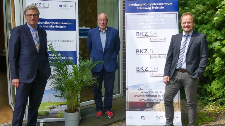 Drei Männer stehen vor einem Gebäude neben Roll-Ups des Breitband-Kompetenzzentrums Schleswig-Holstein. Zwei tragen Anzüge, einer ein Sakko mit Jeans. Eine Pflanze steht im Vordergrund, und im Hintergrund ist eine Tür offen.
