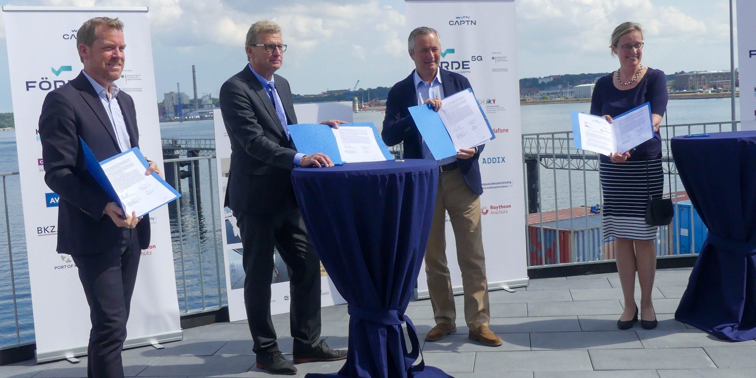 Vier Personen stehen auf einer Terrasse mit Blick auf das Wasser. Sie halten Dokumente und stehen hinter runden Tischen mit blauen Decken. Der Himmel ist bewölkt und im Hintergrund sind Industriebauten zu sehen.