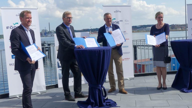 Vier Personen stehen auf einer Terrasse mit Blick auf das Wasser. Sie halten Dokumente und stehen hinter runden Tischen mit blauen Decken. Der Himmel ist bewölkt und im Hintergrund sind Industriebauten zu sehen.