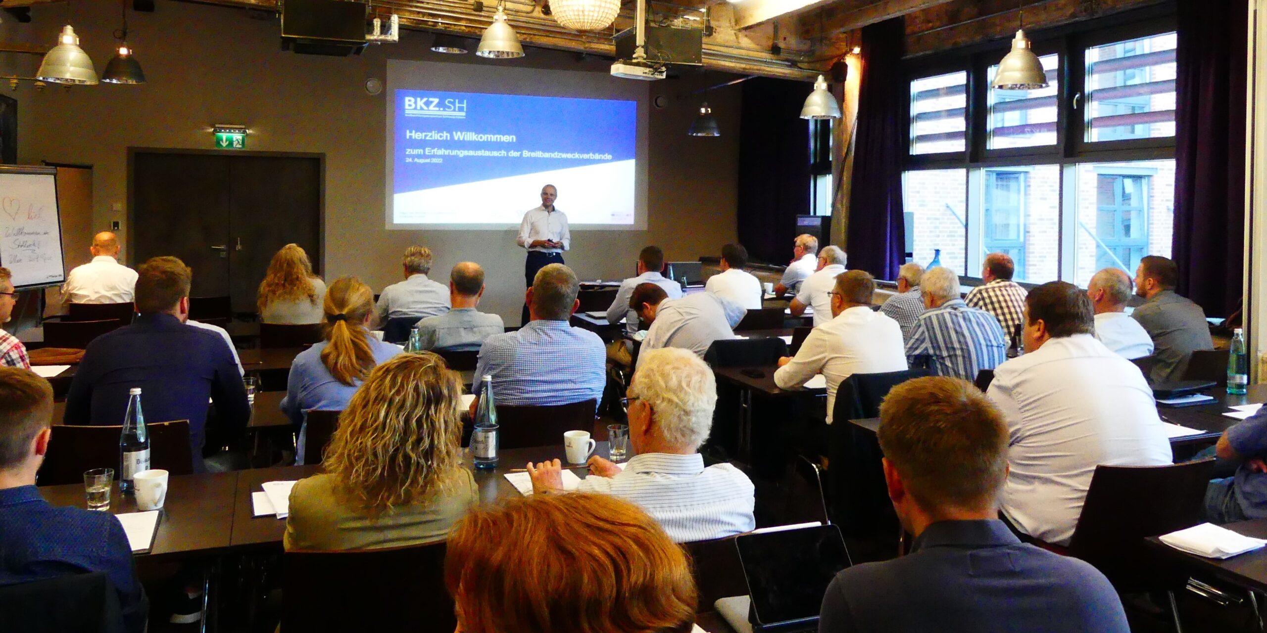 P1020193 Zuschnitt Homepage Ein Konferenzraum mit vielen Personen, die auf Stühlen sitzen und einem Redner zuhören. Der Redner steht vor einer Leinwand, auf der eine Präsentation projiziert wird. Die Wände haben große Fenster und es hängen Lampen von der Decke.