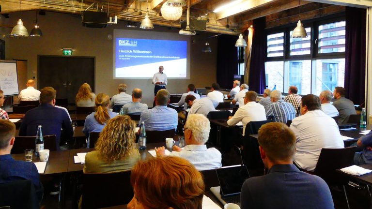 Ein Konferenzraum mit vielen Personen, die auf Stühlen sitzen und einem Redner zuhören. Der Redner steht vor einer Leinwand, auf der eine Präsentation projiziert wird. Die Wände haben große Fenster und es hängen Lampen von der Decke.