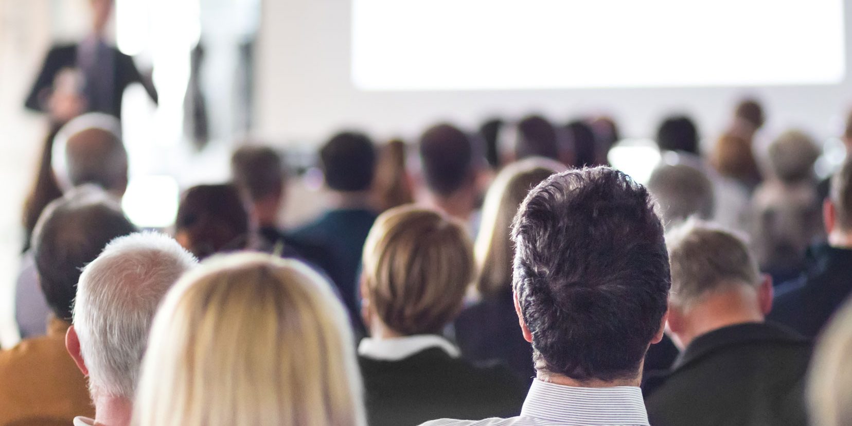 Eine Gruppe von Menschen sitzt in einem Raum und schaut auf eine Leinwand oder Präsentation, die vorne stattfindet. Der Raum ist gut besucht, und die Personen sind von hinten zu sehen.