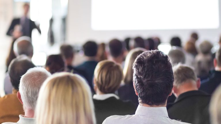 Eine Gruppe von Menschen sitzt in einem Raum und schaut auf eine Leinwand oder Präsentation, die vorne stattfindet. Der Raum ist gut besucht, und die Personen sind von hinten zu sehen.
