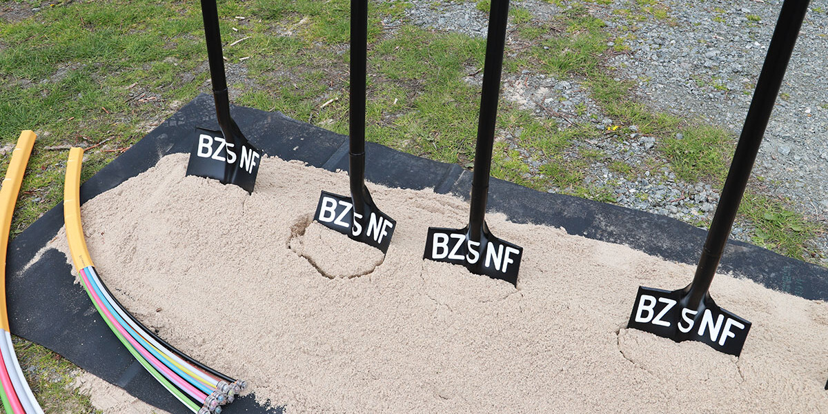 Auf einer Wiese liegen fünf Schaufeln in einer Reihe auf Sand. Jede Schaufel hat die Aufschrift "BZSNF". Rechts am Bildrand ist Schotter, links liegen Rohre mit farbigen Kabeln.