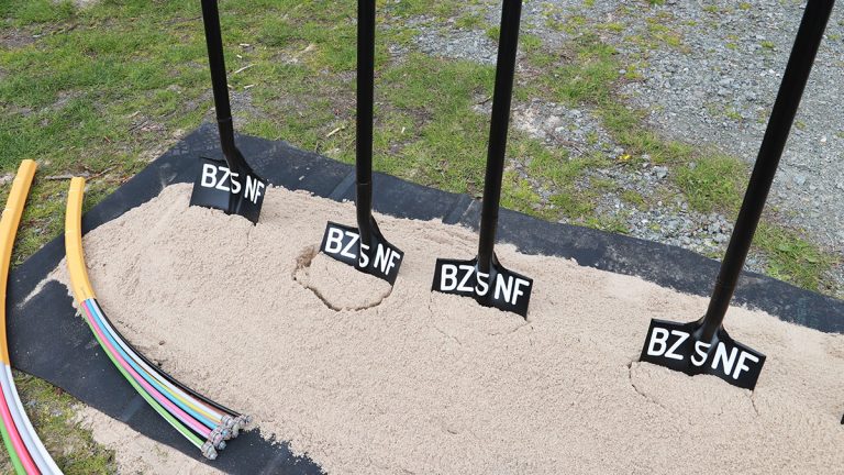 Auf einer Wiese liegen fünf Schaufeln in einer Reihe auf Sand. Jede Schaufel hat die Aufschrift "BZSNF". Rechts am Bildrand ist Schotter, links liegen Rohre mit farbigen Kabeln.