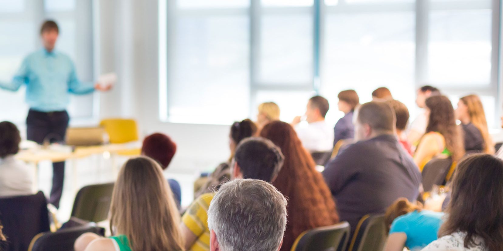 Vortrag Header Ein Vortragssaal mit vielen Menschen, die auf Stühlen sitzen und einem sprechenden Mann vorne zuhören. Der Raum ist durch große Fenster im Hintergrund hell erleuchtet.