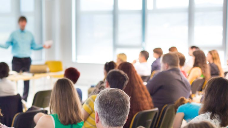 Ein Vortragssaal mit vielen Menschen, die auf Stühlen sitzen und einem sprechenden Mann vorne zuhören. Der Raum ist durch große Fenster im Hintergrund hell erleuchtet.