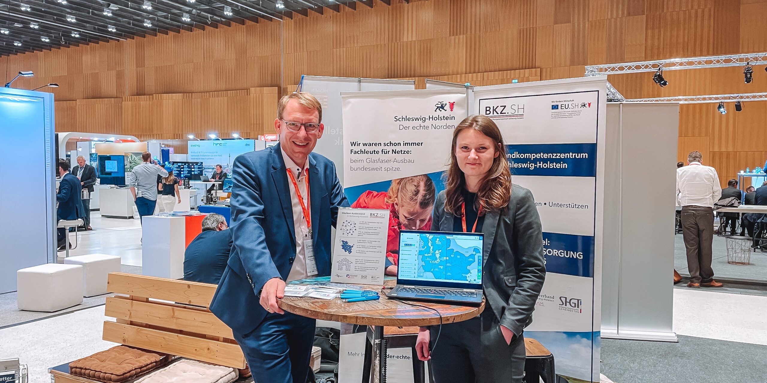 Fiberdays22 Kiara Janpeter Zwei Personen stehen an einem Messestand mit Informationsmaterial und einem Laptop. Im Hintergrund sind weitere Stände sowie eine große Halle mit Holzverkleidung und Deckenbeleuchtung zu sehen.