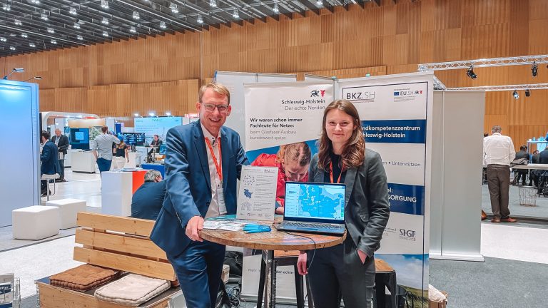 Zwei Personen stehen an einem Messestand mit Informationsmaterial und einem Laptop. Im Hintergrund sind weitere Stände sowie eine große Halle mit Holzverkleidung und Deckenbeleuchtung zu sehen.