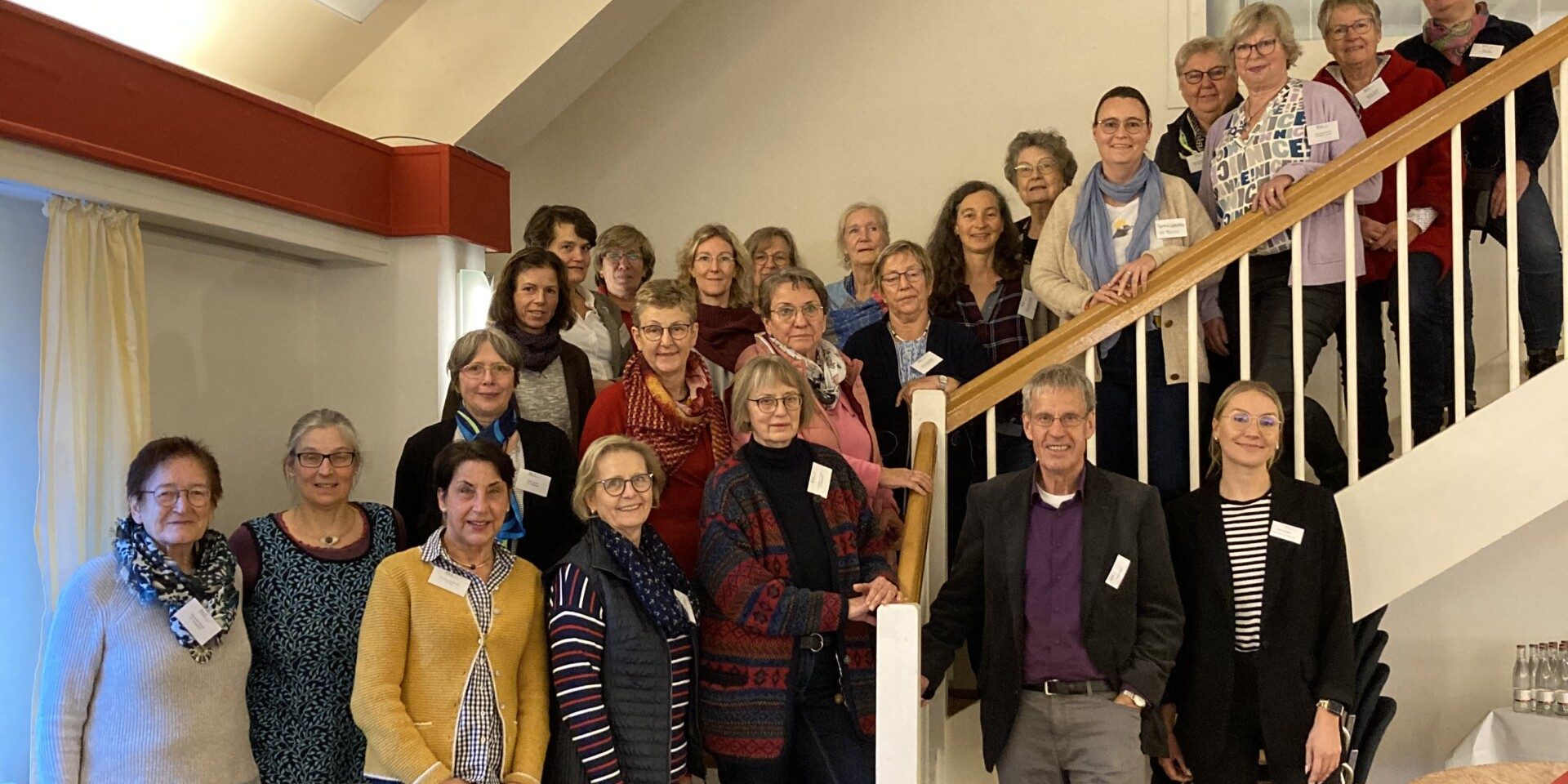 Eine Gruppe von Menschen steht auf einer Treppe und lächelt in die Kamera. Einige Personen stehen auf den Stufen, andere daneben. Der Raum hat Holzboden, und ein großes Fenster ist im Hintergrund zu sehen.