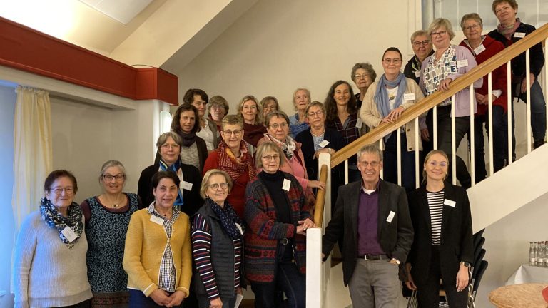 Eine Gruppe von Menschen steht auf einer Treppe und lächelt in die Kamera. Einige Personen stehen auf den Stufen, andere daneben. Der Raum hat Holzboden, und ein großes Fenster ist im Hintergrund zu sehen.
