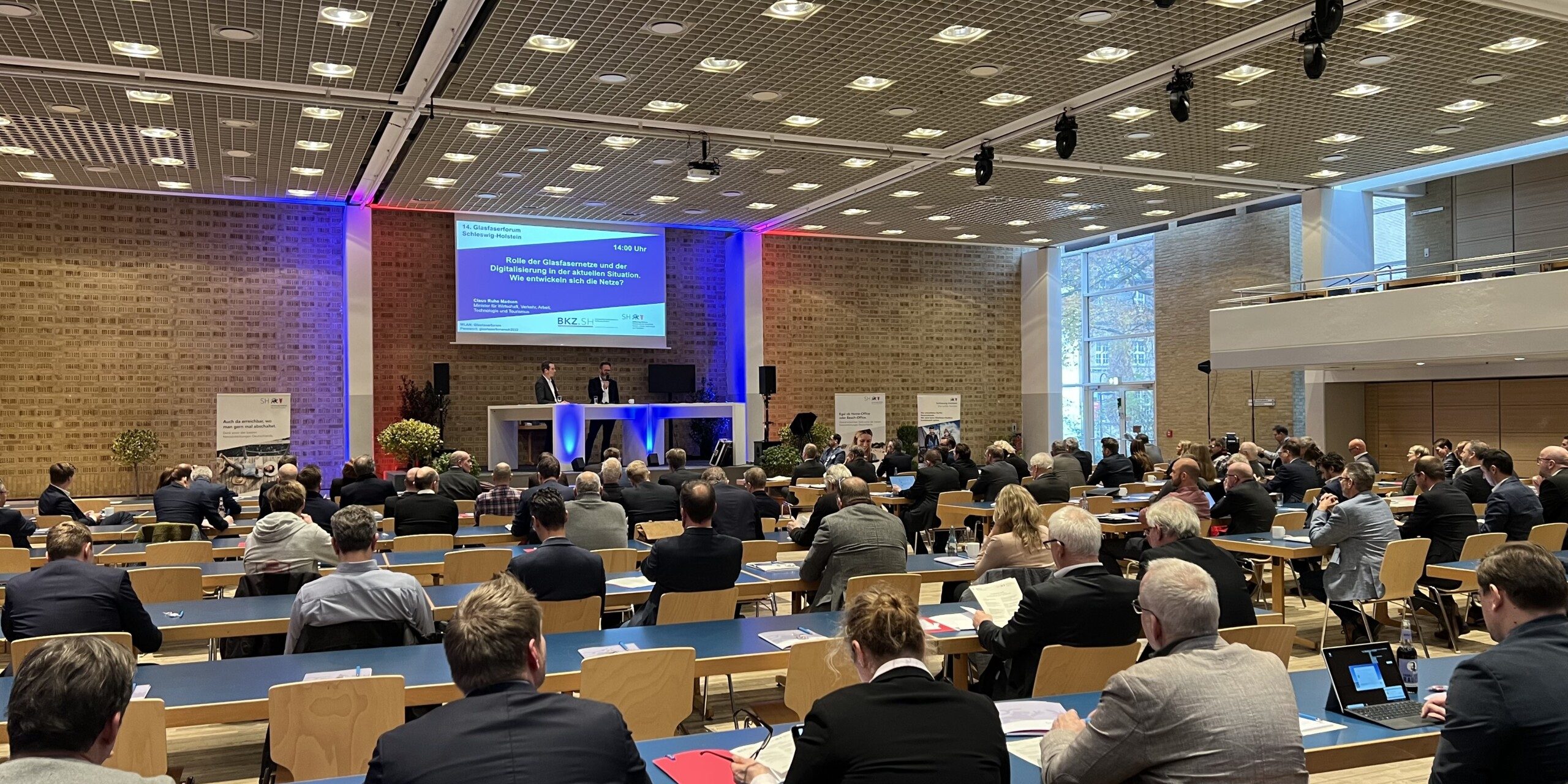 Image00006 In einem großen Konferenzraum sitzen zahlreiche Personen an Tischen und hören zwei Rednern auf einer erhöhten Bühne zu. Der Raum hat eine hohe Decke mit vielen Lichtern und große Fenster auf der rechten Seite.
