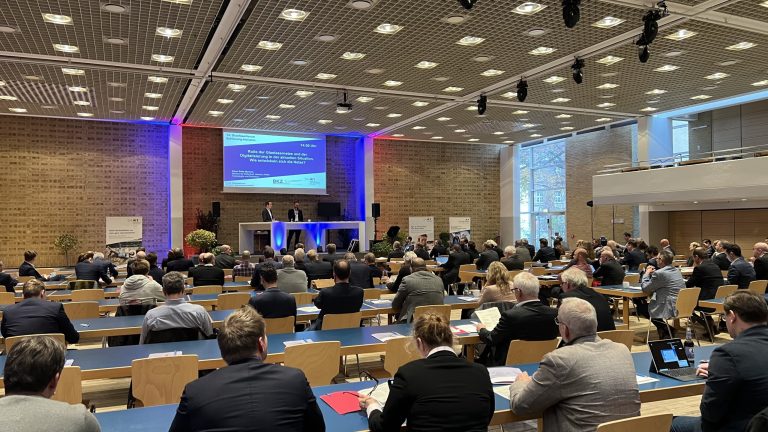 In einem großen Konferenzraum sitzen zahlreiche Personen an Tischen und hören zwei Rednern auf einer erhöhten Bühne zu. Der Raum hat eine hohe Decke mit vielen Lichtern und große Fenster auf der rechten Seite.