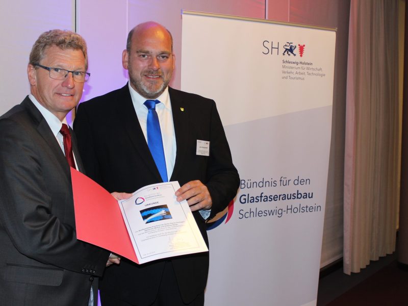 Zwei Männer stehen nebeneinander und halten gemeinsam ein aufgeschlagenes Dokument. Hinter ihnen befindet sich ein Roll-up mit der Aufschrift "Bündnis für den Glasfaserausbau Schleswig-Holstein".