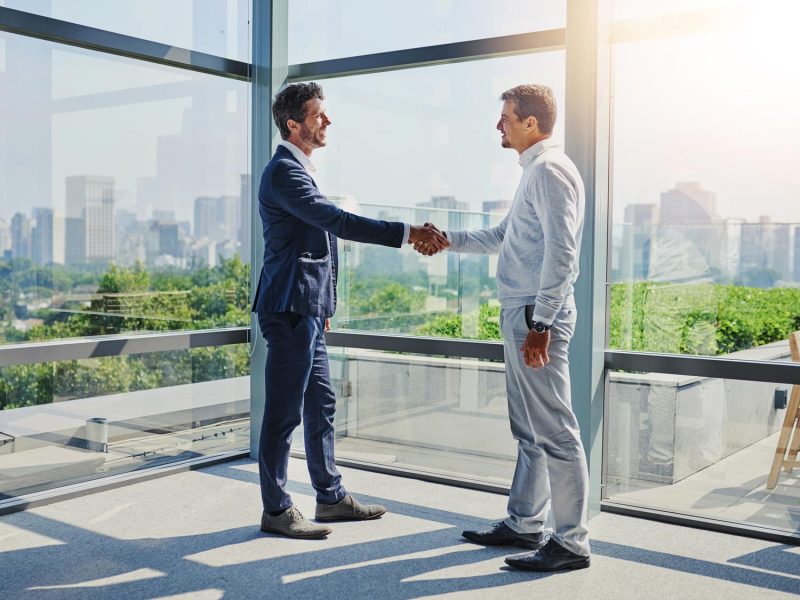 Zwei Männer stehen in einem modernen Büro mit großen Fenstern, die eine Stadt und Bäume im Hintergrund zeigen. Sie geben sich die Hand, während Sonnenlicht den Raum erhellt.