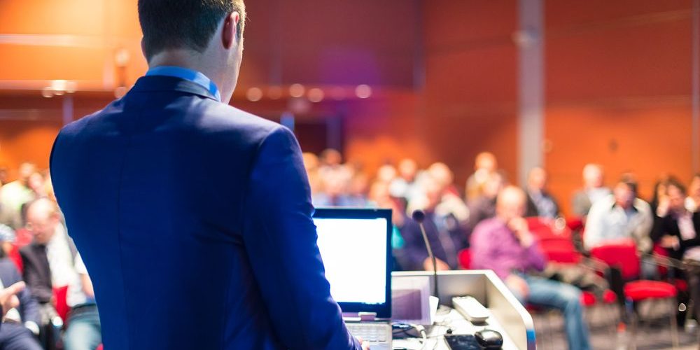 Shutterstock 196793828 Ein Mann in einem Anzug steht an einem Rednerpult mit einem Laptop, während er zu einem Publikum in einem Konferenzraum spricht. Die Zuhörer sitzen auf roten Stühlen und der Raum ist orange beleuchtet.