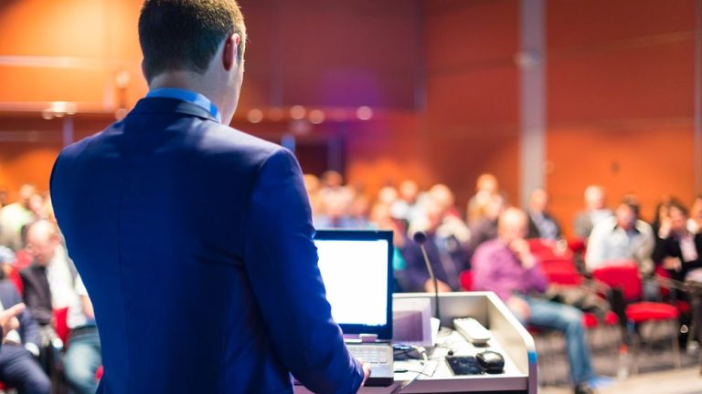 Ein Mann in einem Anzug steht an einem Rednerpult mit einem Laptop, während er zu einem Publikum in einem Konferenzraum spricht. Die Zuhörer sitzen auf roten Stühlen und der Raum ist orange beleuchtet.