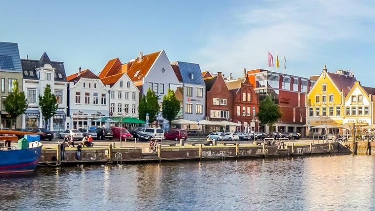 Panorama eines malerischen Hafens mit bunten, traditionellen Häusern am Wasser. Im Vordergrund liegt ein großes blaues Boot. Die Szene ist von einem klaren, blauen Himmel beleuchtet.