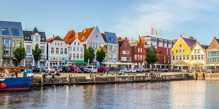 Slider Panorama eines malerischen Hafens mit bunten, traditionellen Häusern am Wasser. Im Vordergrund liegt ein großes blaues Boot. Die Szene ist von einem klaren, blauen Himmel beleuchtet.